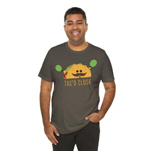 Könnte beinhalten: Dunkelgraues T-Shirt mit einem Cartoon-Taco mit Schnurrbart und grünen Hanteln. Der Text "TAC'O CLOCK" ist darunter aufgedruckt. Das T-Shirt hat einen lässigen Stil.