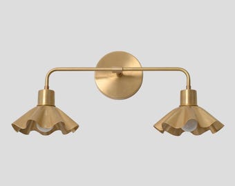 May include: A two-light wall sconce featuring a circular brass backplate and a horizontal bar supporting two downward-facing, ruffled-edge brass shades. Each shade houses a single light bulb, creating a symmetrical fixture with a metallic finish.
