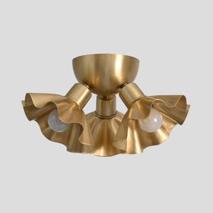 Può includere: Un lampadario da soffitto color ottone con tre paralumi a balze, ciascuno con una lampadina. L'apparecchio ha una base centrale a forma di ciotola e tre bracci regolabili. Il design è elegante e moderno.