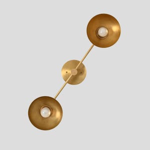 May include: A wall-mounted light fixture featuring a brushed brass finish with a central circular mounting plate and two adjustable arms extending diagonally. Each arm terminates in a wide, bowl-shaped shade containing a single white light bulb.
