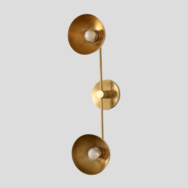 May include: A wall-mounted light fixture featuring a vertical brass-toned metal rod with a circular mounting plate in the center. Two conical shades extend from the rod, each housing a single round white light bulb facing outward from the fixture.
