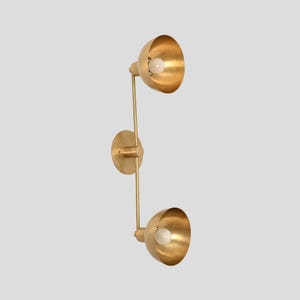 May include: A wall-mounted light fixture with a brushed brass finish, featuring a central circular mounting plate and a vertical arm that supports two adjustable, bowl-shaped shades, each containing a single round white light bulb.