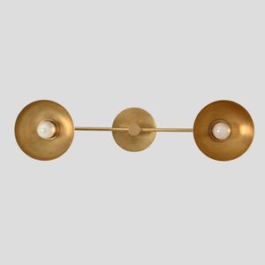 May include: A horizontal wall light fixture featuring a central circular mounting plate and a thin metal bar extending to two circular, bowl-shaped shades in a brushed brass finish, each housing a single white globe light bulb in the center.