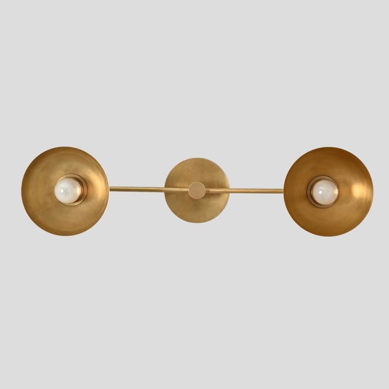 May include: A horizontal wall light fixture featuring a central circular mounting plate and a thin metal bar extending to two circular, bowl-shaped shades in a brushed brass finish, each housing a single white globe light bulb in the center.