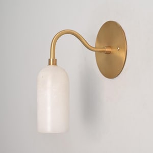May include: A wall sconce featuring a circular brass backplate and a curved brass arm that supports a cylindrical white stone shade. The fixture has a metallic gold finish and a smooth, opaque white stone texture on the downward-facing light cover.