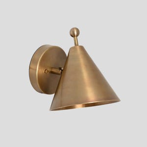May include: A brass wall sconce with a cone-shaped shade. The sconce has a simple, modern design.