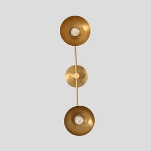 May include: A vertical wall sconce featuring a brushed brass finish with two circular, bowl-shaped shades. A central circular mounting plate connects the two arms, each holding a single white light bulb centered within the metallic gold-toned bowls.
