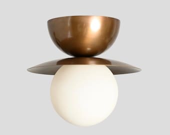 May include: A ceiling light fixture featuring a large, matte bronze-colored metal bowl at the top, a flat circular bronze disc in the middle, and a smooth, spherical white glass globe at the bottom, creating a layered, geometric lighting design.