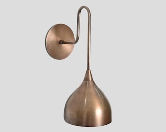 May include: A wall-mounted lamp featuring a circular backplate and a curved arm that supports a downward-facing, bell-shaped shade. The entire fixture is finished in a metallic bronze tone with a smooth, polished surface throughout the design.