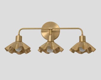 May include: A three-light wall fixture in a brushed brass finish featuring a circular mounting plate and a horizontal bar. Each of the three lights has a downward-facing, ruffled, bell-shaped shade with a white bulb visible in the center.