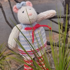 Può includere: Un animale di peluche lavorato a maglia a forma di orso che indossa un vestito a righe blu e bianche con un nastro rosso. L'orso è seduto su un ramo di un pino.