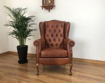 May include: A brown leather wingback chair featuring a tufted backrest with button detailing, rolled arms with brass nailhead trim, and curved wooden legs. The chair has a high back and a single seat cushion, providing a structured seating option.