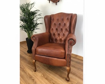 May include: A brown leather armchair with a high, button-tufted back and rolled arms. The chair has wooden legs and is set against a white wall. A potted plant is visible.