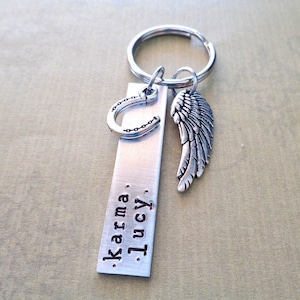 May include: Silver keyring with a horseshoe charm, an angel wing charm, and a rectangular tag engraved with the text "Karma. Lucy."