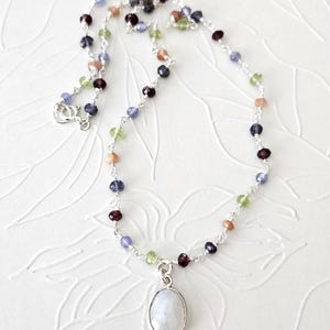 May include: A delicate silver chain necklace featuring a series of small, multi-colored faceted gemstone beads in shades of blue, green, red, and peach. The necklace includes a single oval-shaped, iridescent moonstone pendant at the center.