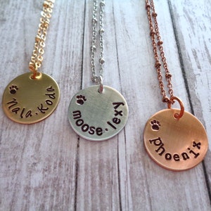 May include: Three gold, silver and copper necklaces with round pendants. Each pendant has a paw print and a name engraved on it. The names are "Mia Koda", "Moose Lexy", and "Phoenix".