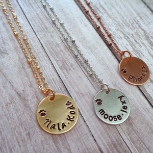 May include: Three gold, silver and copper necklaces with round pendants. Each pendant has a paw print and a name engraved on it. The names are "Nala.koba", "Moose.Lex", and "Phoenix".