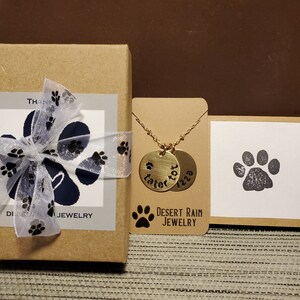 May include: A gold necklace with two round pendants engraved with the names "Tater Tot" and "Izzy". The necklace is packaged in a brown box with a white label that says "Desert Rain Jewellery".