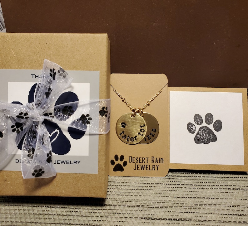 May include: A gold necklace with two round pendants engraved with the names "Tater Tot" and "Izzy". The necklace is packaged in a brown box with a white label that says "Desert Rain Jewellery".