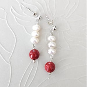 May include: A pair of drop earrings featuring three white freshwater pearls stacked vertically above a single round red bead, all suspended from silver-toned metal posts with matching butterfly backings for secure attachment to the earlobe.