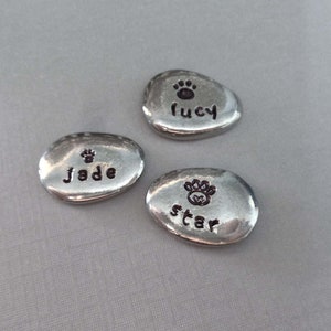 May include: Three silver-coloured, oval-shaped, personalised pet memorial stones with a paw print and the names "Lucy", "Jade", and "Star" engraved on them.