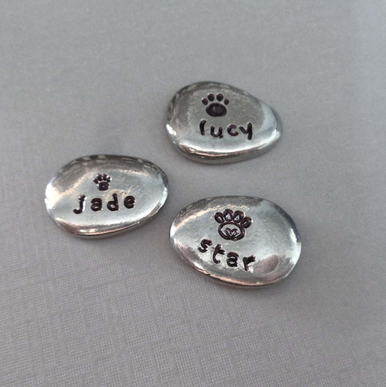 May include: Three silver-coloured, oval-shaped, personalised pet memorial stones with a paw print and the names "Lucy", "Jade", and "Star" engraved on them.