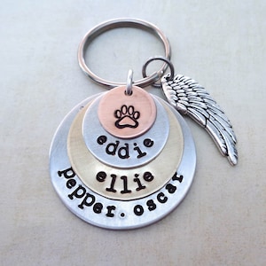 May include: A silver key chain with four stacked metal discs. The top disc is copper and features a paw print. The other discs are silver and have the names "pepper", "ellie", "eddie", and "oscar" engraved on them. A silver angel wing charm hangs from the top of the key chain.
