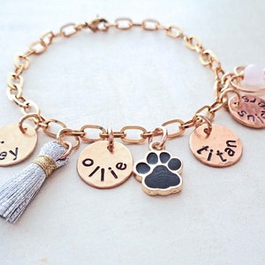 May include: Gold chain bracelet with multiple charms, including a paw print, a tassel, and three round charms with the names "Joey", "Ollie", and "Titan" engraved on them.