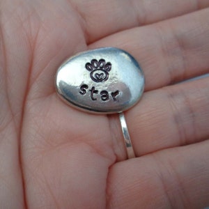 May include: A silver-coloured, oval-shaped charm with a paw print and the word "star" engraved on it.