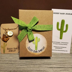 May include: A brown cardboard box with a green bow and a sticker that says "Desert Rain Jewelry" and a cactus illustration. The box is sitting on a brown surface. A business card with the text "Desert Rain Jewelry" and a cactus illustration is visible in the background.