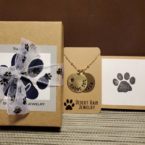 May include: A gold necklace with two round pendants that say "Tater Tot" and "Izzy" on them. The necklace is displayed on a brown card with a paw print and the text "Desert Rain Jewellery".
