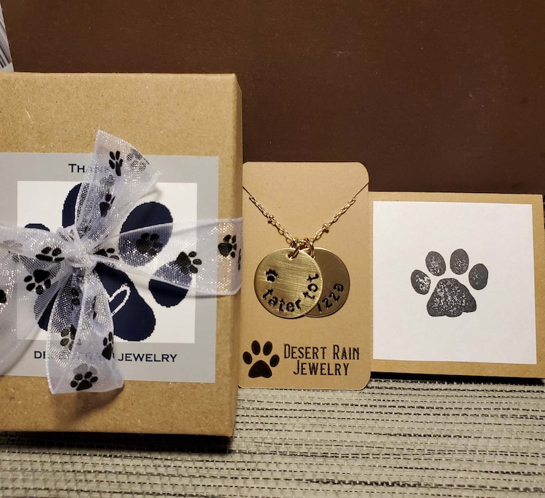 May include: A gold necklace with two round pendants that say "Tater Tot" and "Izzy" on them. The necklace is displayed on a brown card with a paw print and the text "Desert Rain Jewellery".
