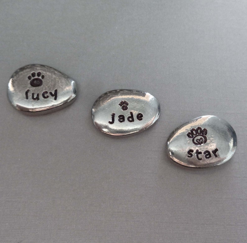 May include: Three silver oval-shaped pet memorial stones with paw prints and the names Lucy, Jade, and Star engraved on them.