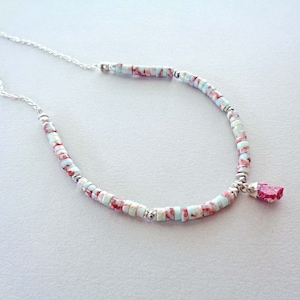 Op de afbeelding: Een delicate ketting met een zilveren ketting en kleine, lichtblauwe en rode kralen. Het heeft een kleine, roze edelsteen hanger. De kralen zijn gelijkmatig verdeeld, waardoor een subtiel, elegant ontwerp ontstaat.