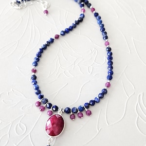 May include: A necklace featuring faceted dark blue lapis lazuli beads interspersed with small magenta ruby beads. A large, oval, faceted magenta ruby pendant is set in a silver frame at the center, accented by small dangling ruby beads and a single blue bead.