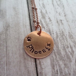 May include: A rose gold necklace with a round pendant that says "phoenix" with a paw print above the word.