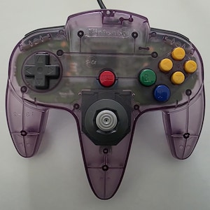 May include: A transparent purple Nintendo 64 controller. The controller features a gray directional pad, a red A button, a green B button, a blue Z button, and yellow C buttons. The controller has a black analog stick and the Nintendo logo.