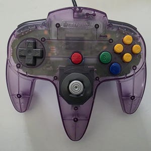 May include: A translucent purple Nintendo 64 controller with a black directional pad, yellow, green, and blue buttons, and a red 'Start' button. The controller has a central analog stick and a Nintendo logo.