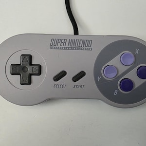 May include: A Super Nintendo Entertainment System controller in light gray with a black directional pad, select and start buttons, and purple X, Y, A, and B buttons. The controller has the words "SUPER NINTENDO ENTERTAINMENT SYSTEM" printed on it.