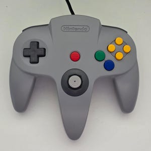 May include: A gray Nintendo 64 video game controller featuring a black directional pad, a central red start button, a gray analog stick, and four colored action buttons in yellow, green, and blue, with a black cord extending from the top.