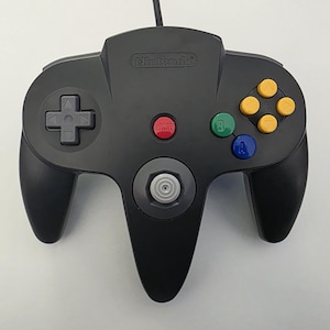 May include: Black Nintendo 64 controller with a gray directional pad, yellow, green, blue, and red buttons. The controller has a three-pronged design and the Nintendo logo is visible.