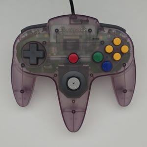 May include: A translucent purple video game controller featuring a central gray analog stick, a black directional pad, a red start button, and four colored action buttons in yellow, green, and blue. The Nintendo logo is embossed at the top center.