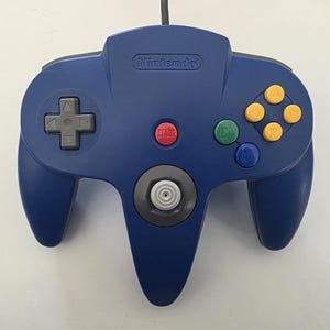 May include: A blue Nintendo 64 controller with a gray directional pad, yellow action buttons, and a red start button. The word "Nintendo" is printed at the top. The controller has two handles and a central analog stick.