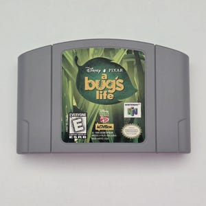 May include: A gray Nintendo 64 video game cartridge for A Bug's Life. The label features a green leaf background with the title text, Disney Pixar logo, ESRB E rating, Nintendo 64 logo, and Activision branding. The cartridge is rectangular with rounded top corners.