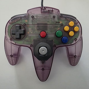 May include: A transparent purple Nintendo 64 controller with a black directional pad, yellow, green, and blue buttons, and a red start button. The controller has a black cord and is set against a white background.