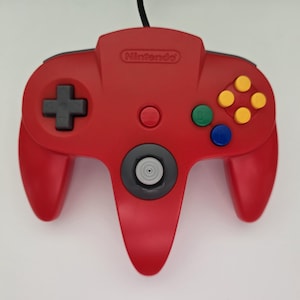 May include: A red Nintendo 64 video game controller featuring a gray analog stick, a black directional pad, four yellow C-buttons, a green C-button, a blue C-button, and a red Start button. The Nintendo logo is embossed at the top center.