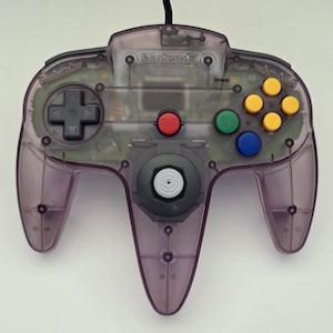 May include: A translucent purple video game controller featuring a central gray analog stick, a black directional pad, a red start button, a green B button, a blue A button, and four yellow C buttons. The Nintendo logo is embossed at the top center.