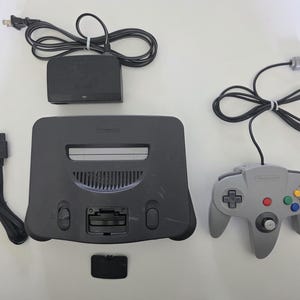 May include: A complete Nintendo 64 gaming console set, featuring a gray console, a gray controller with colored buttons, a power adapter, and AV cables. The set includes a black expansion pack cover.