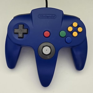 May include: A blue Nintendo 64 video game controller featuring a central gray analog stick, a black directional pad on the left, four yellow C-buttons, a green B-button, a blue A-button, and a red Start button centered on the controller body.