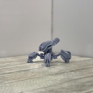 Puede incluir: Un modelo de robot con forma de araña de color gris, impreso en 3D, con un cañón grande en la parte superior. El robot tiene seis patas y está en posición de caminar.